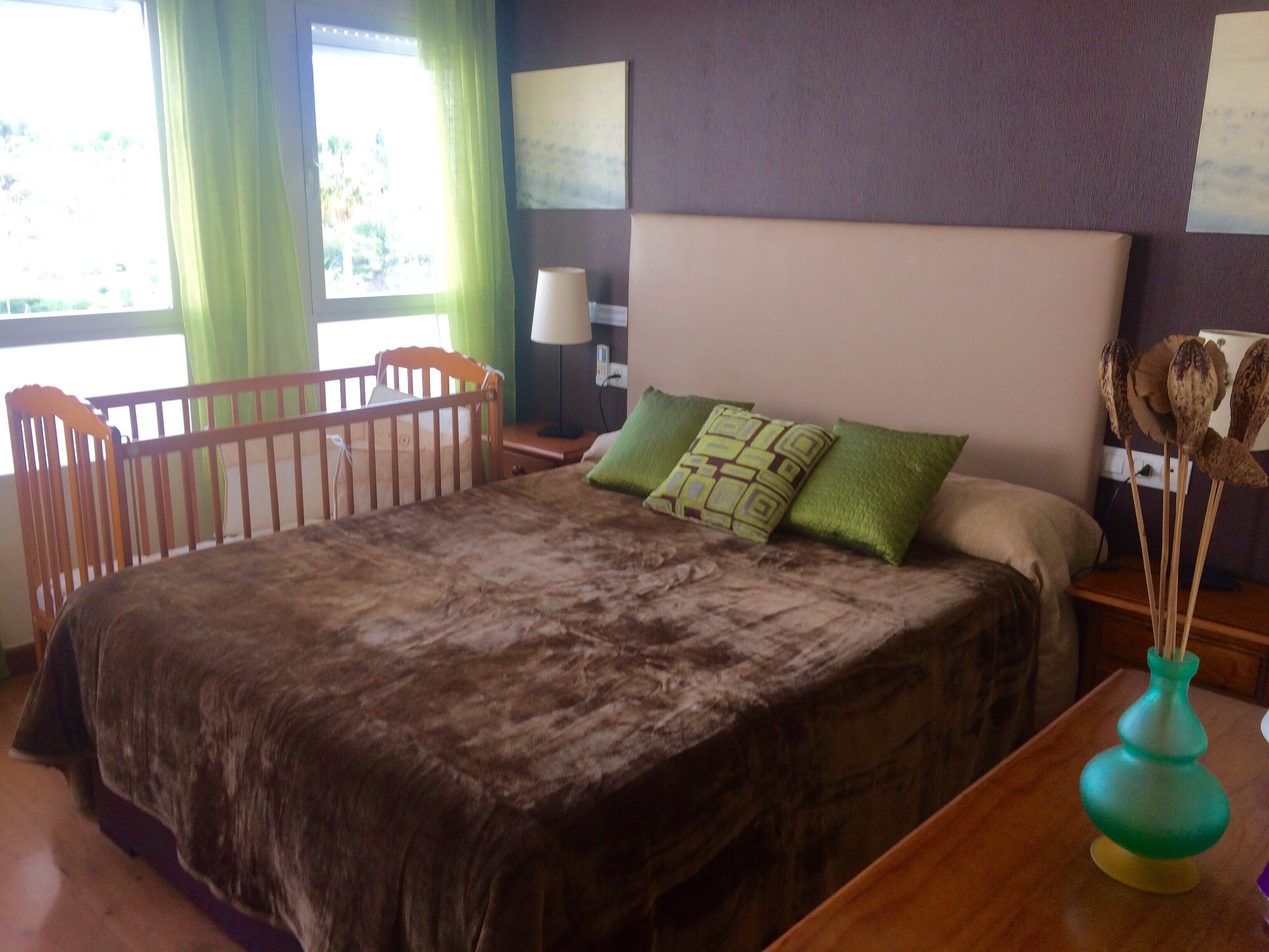 A bedroom with a large bed, a wooden crib, a nightstand, a lamp, and a vase with dried flowers.