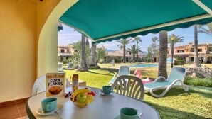 Una mesa con un bol de fruta, una caja de copos de maíz y dos tazas, ubicada bajo un toldo verde con vistas a una piscina y edificios de un complejo turístico.