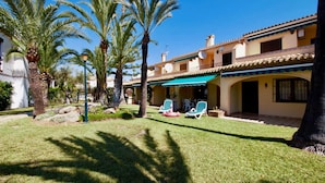 Propiedad con césped cuidado, palmeras y zona de estar exterior con sillones de descanso.