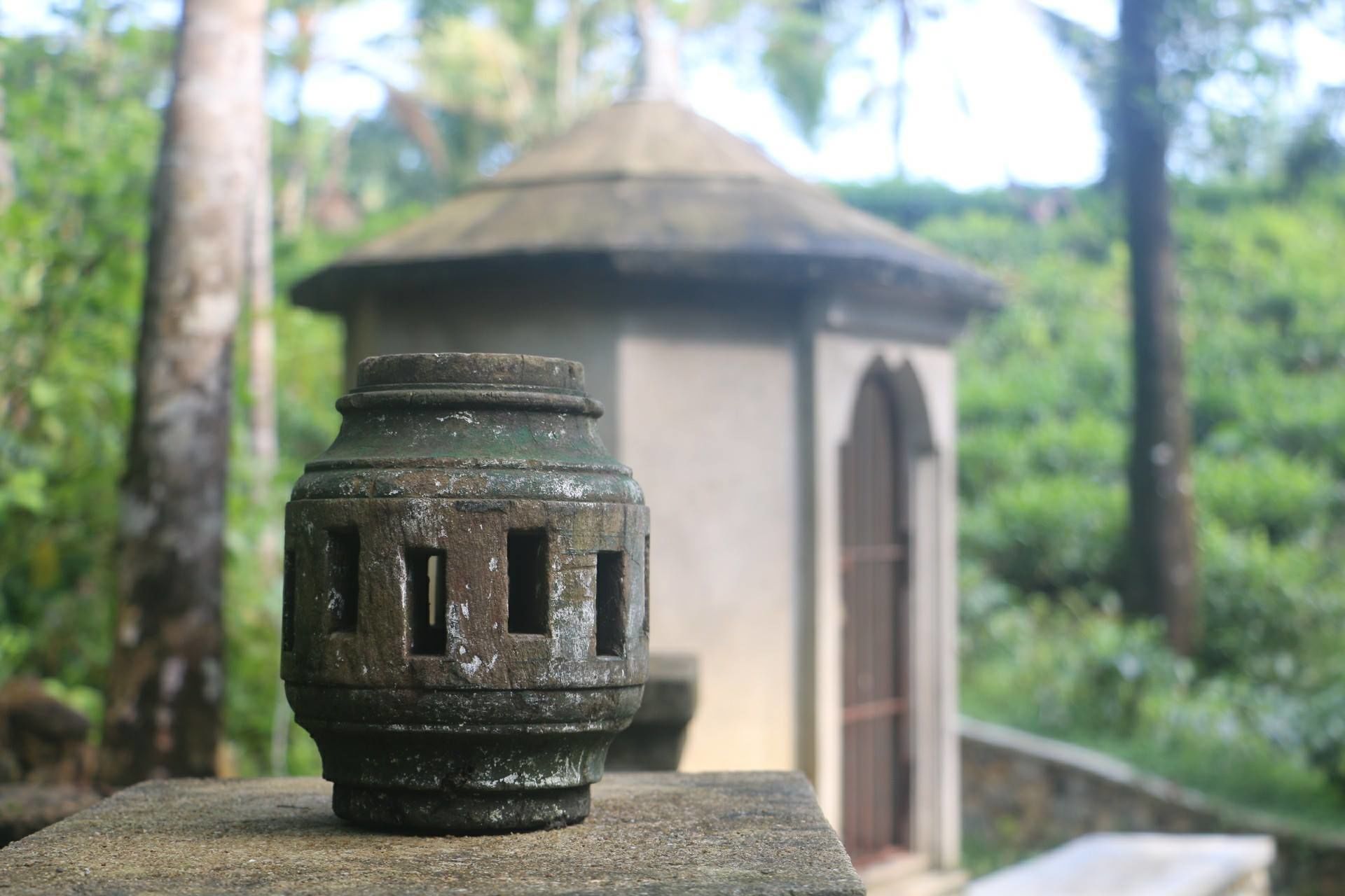A stone lantern with a weathered appearance, set against a backdrop of a traditional building with a tiled roof, surrounded by lush greenery.