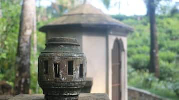 A stone lantern with a weathered appearance, set against a backdrop of a traditional building with a tiled roof, surrounded by lush greenery.