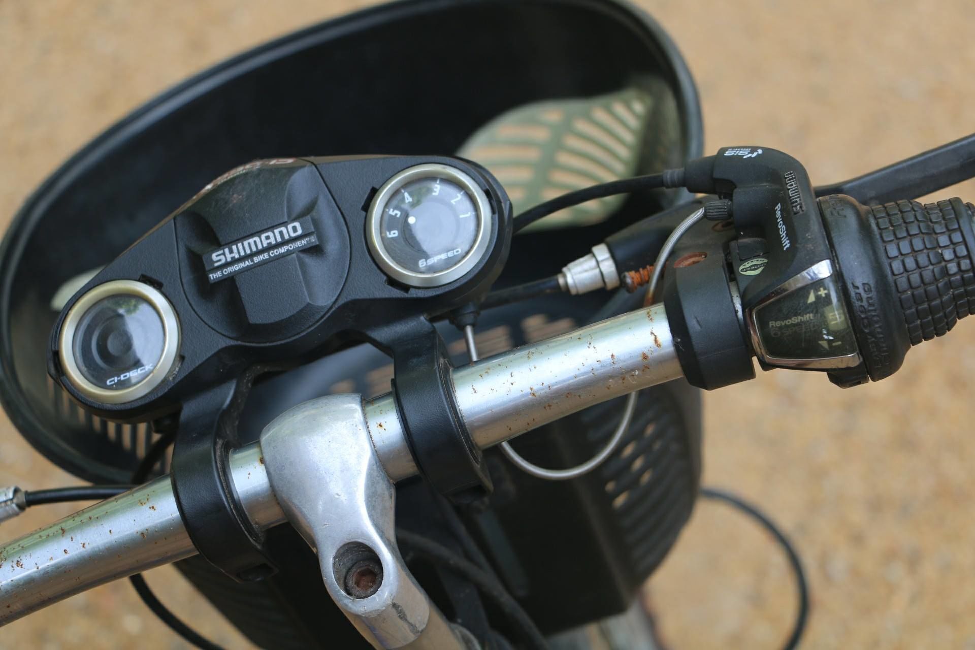 A close-up of a motorcycle's handlebar with a speedometer, a Shimano brand component, and a cable.