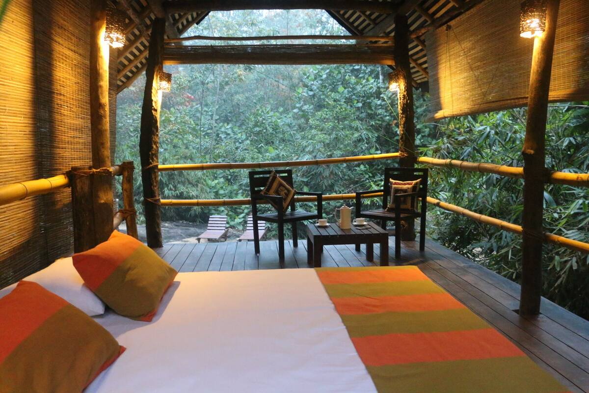 A room with a bed, two chairs, and a table, all made of wood, with a view of dense greenery outside.