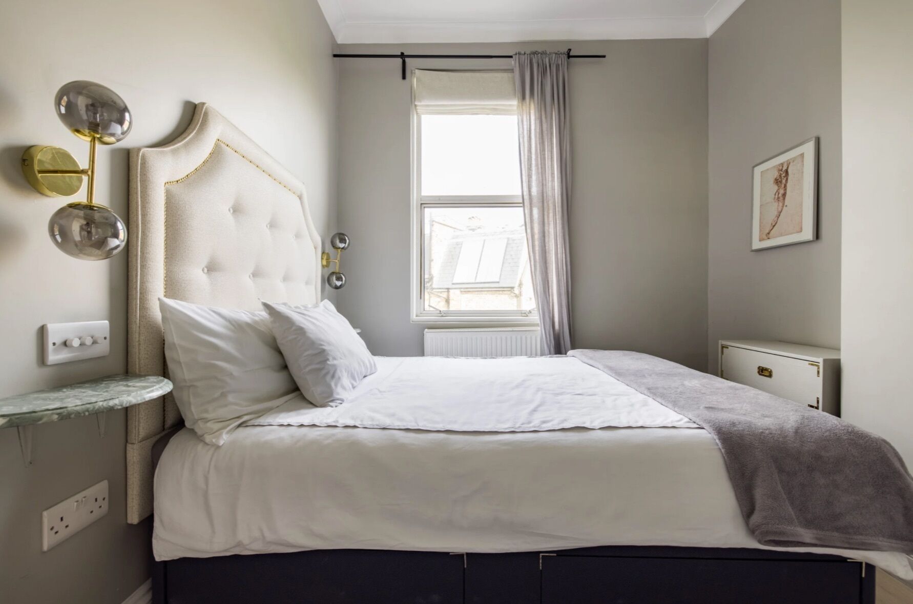 Une chambre à coucher comprenant un lit, une table de chevet, une fenêtre avec des rideaux et un luminaire fixé au mur.