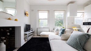 A modern living room with a white sofa, black rug, and a TV on a white stand. There are windows with white frames and a ceiling light fixture. - Elegant apartment in Kensington Olympia (available for LONGER LETS) (London)