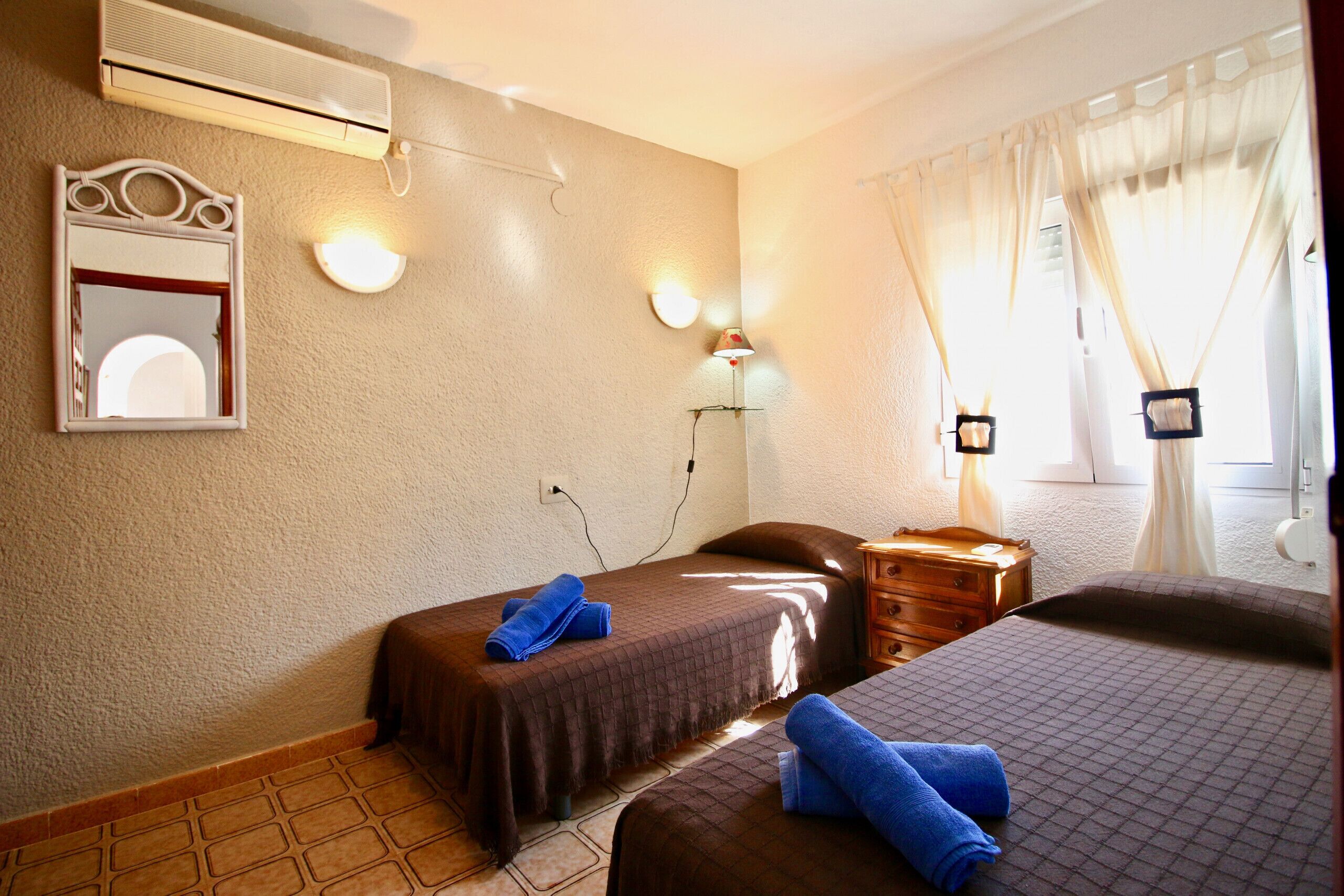 A hotel room with two beds, a mirror, wall-mounted lights, an air conditioner, and a window with curtains.