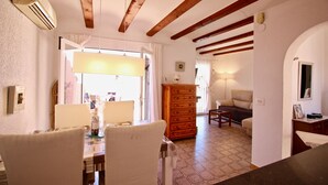 Un área de comedor con una mesa y sillas, un armario de madera y un sofá en una habitación con vigas de madera a la vista y suelo de baldosas.