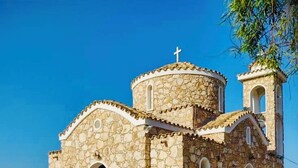 A stone church with a bell tower and a cross on top, located on a paved area with a clear view of the sea in the background.