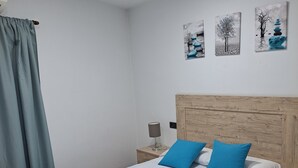 Una cama bien hecha con sábanas blancas y almohadas azules, un cabecero de madera, una mesita de noche con una lámpara y tres obras de arte en la pared que representan árboles.
