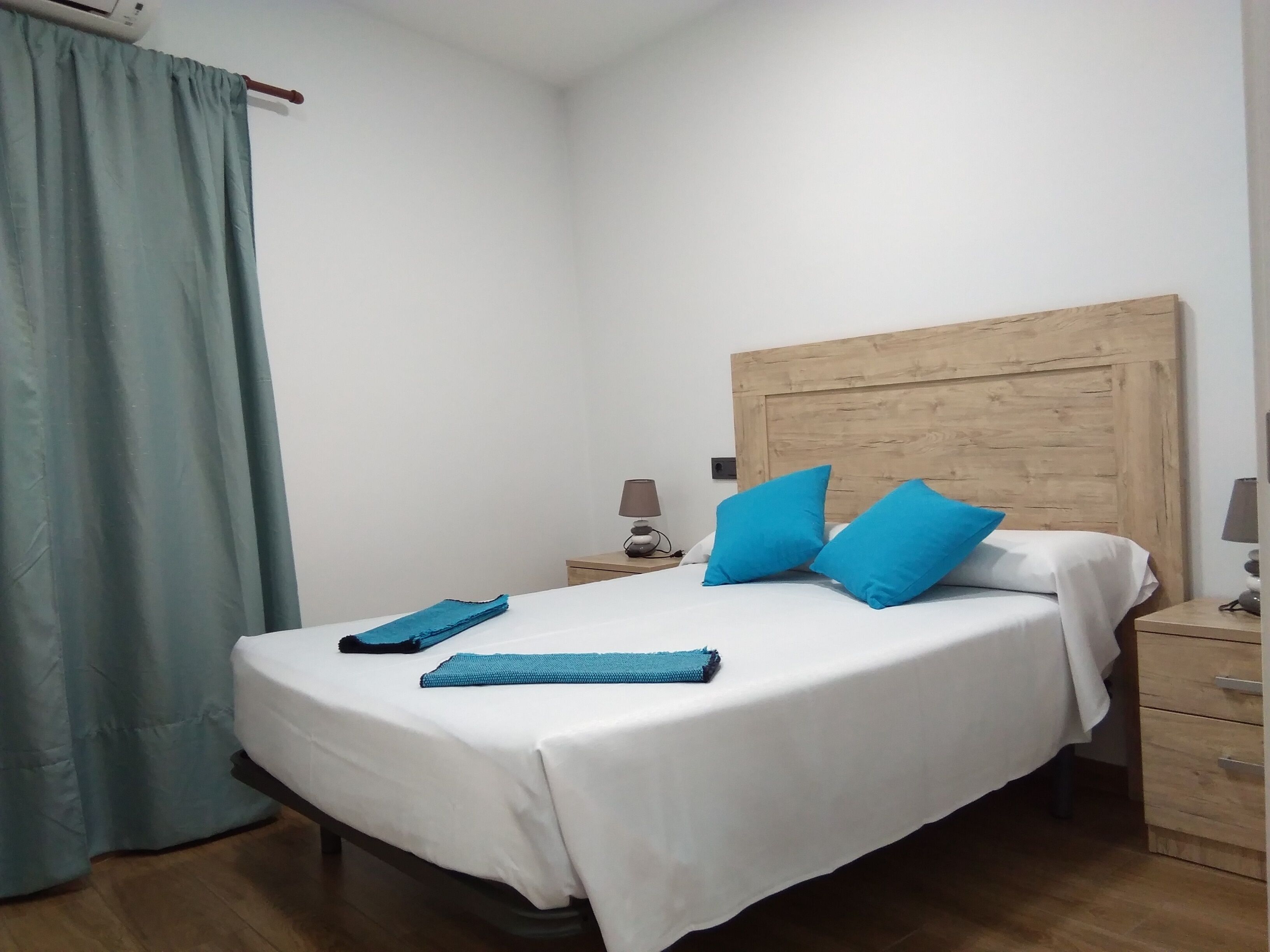 A neatly made bed with white sheets and two blue pillows, a wooden headboard, a bedside table with a lamp, and a wooden floor.