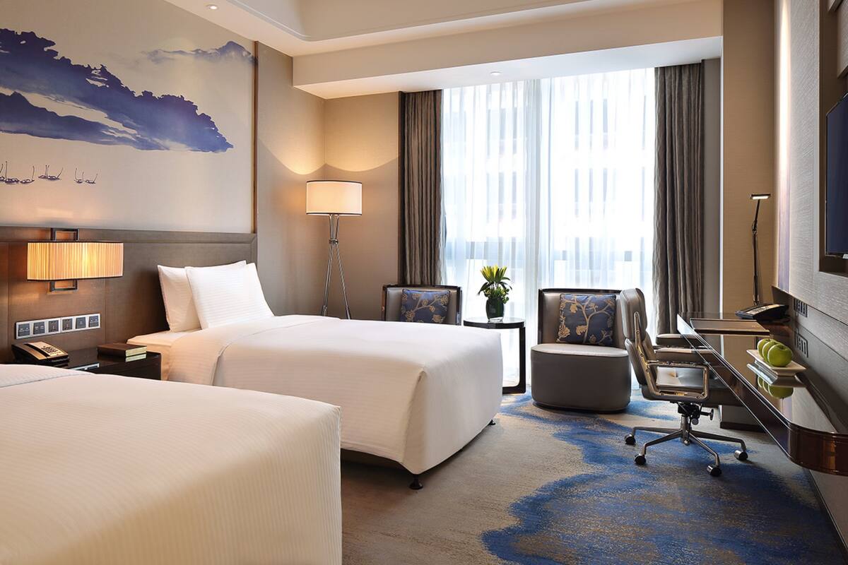 A hotel room with two beds, a desk with a chair, a lamp, a TV, and a large window with curtains.