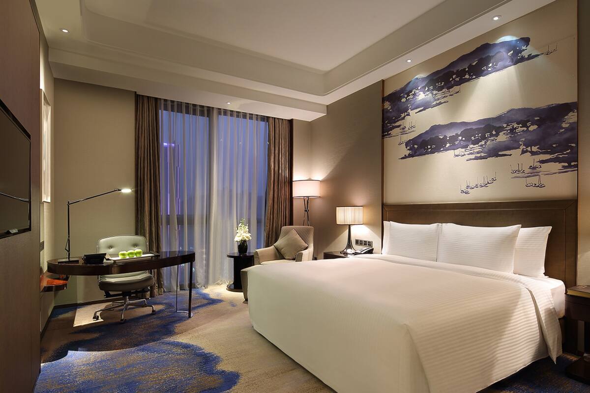A hotel room with a large bed, a desk with a chair, a nightstand with a lamp, and a large window with curtains.