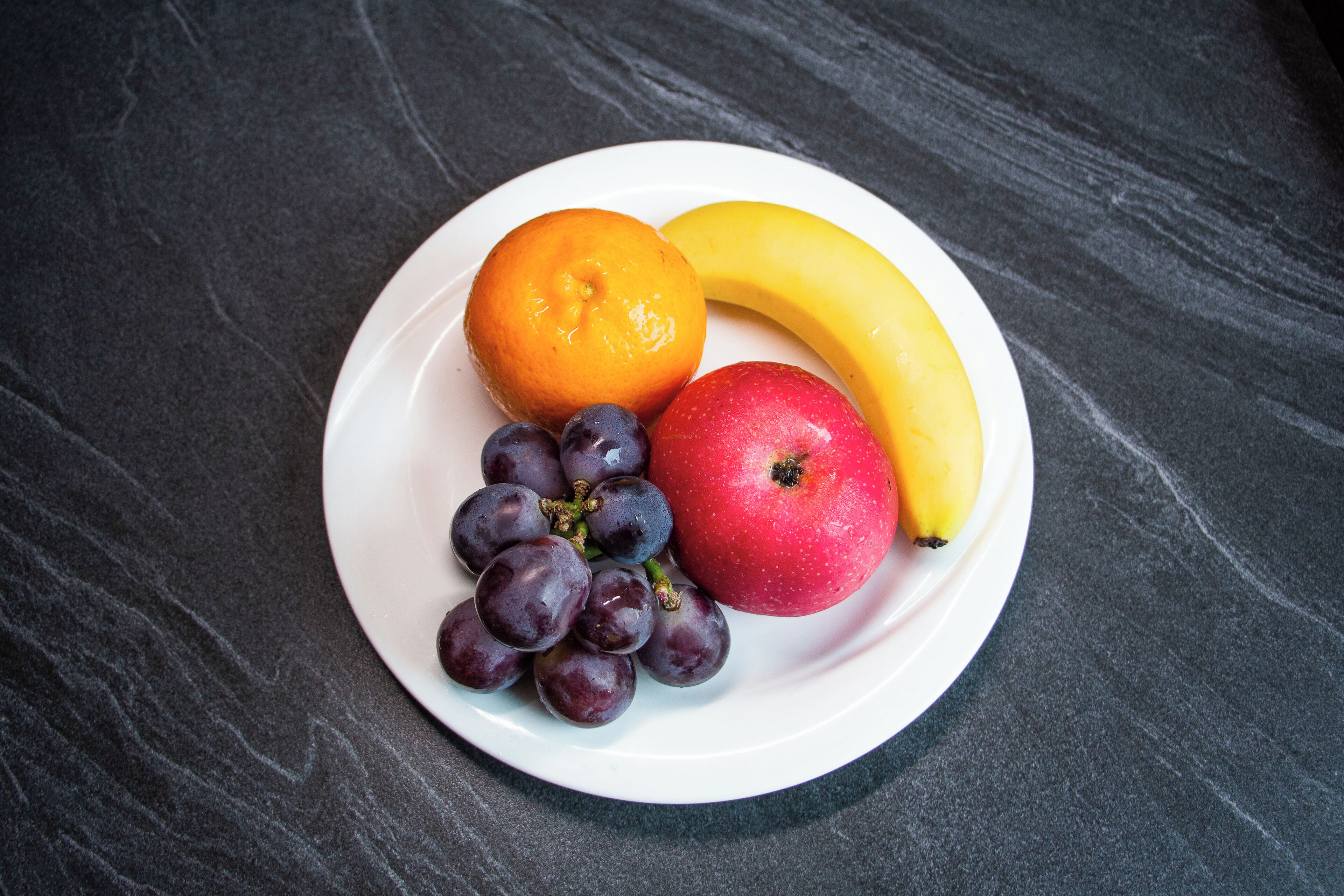 A plate with an orange, a banana, and a red apple on a dark surface.