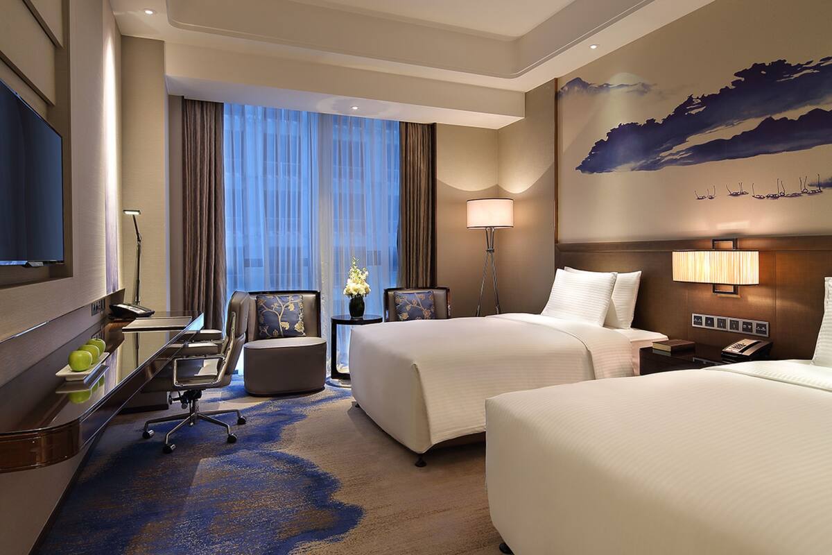 A hotel room with two beds, a desk with a chair, a flat-screen TV, a lamp, and a painting on the wall.
