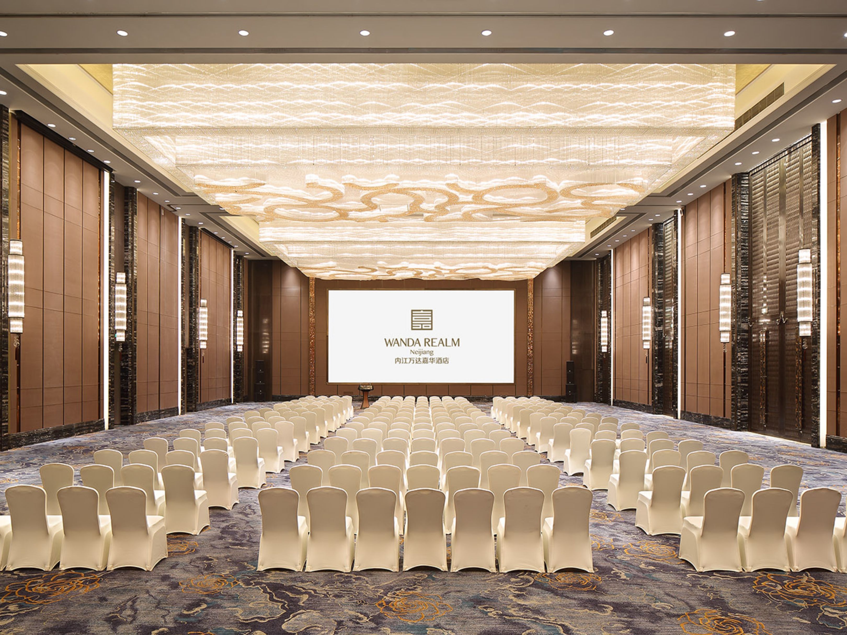 A conference room with rows of chairs, a large screen displaying 'Wanda Realm', and a patterned ceiling.