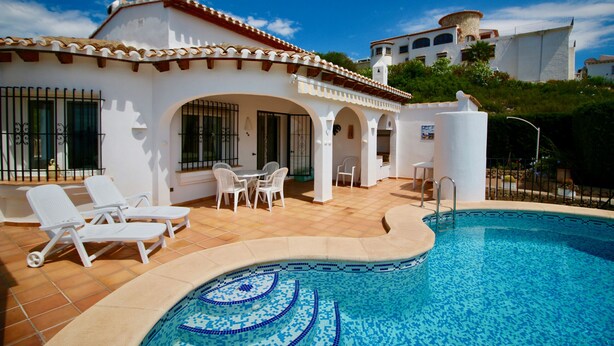 Un área junto a la piscina con sillas de descanso, una piscina con mosaicos azules y un edificio blanco con arcos y techo de tejas.