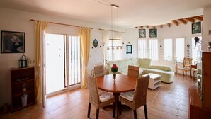 Un amplio espacio de estar con una mesa redonda de madera, sillas de mimbre, un sofá de color claro y un comedor con una mesa pequeña y sillas.