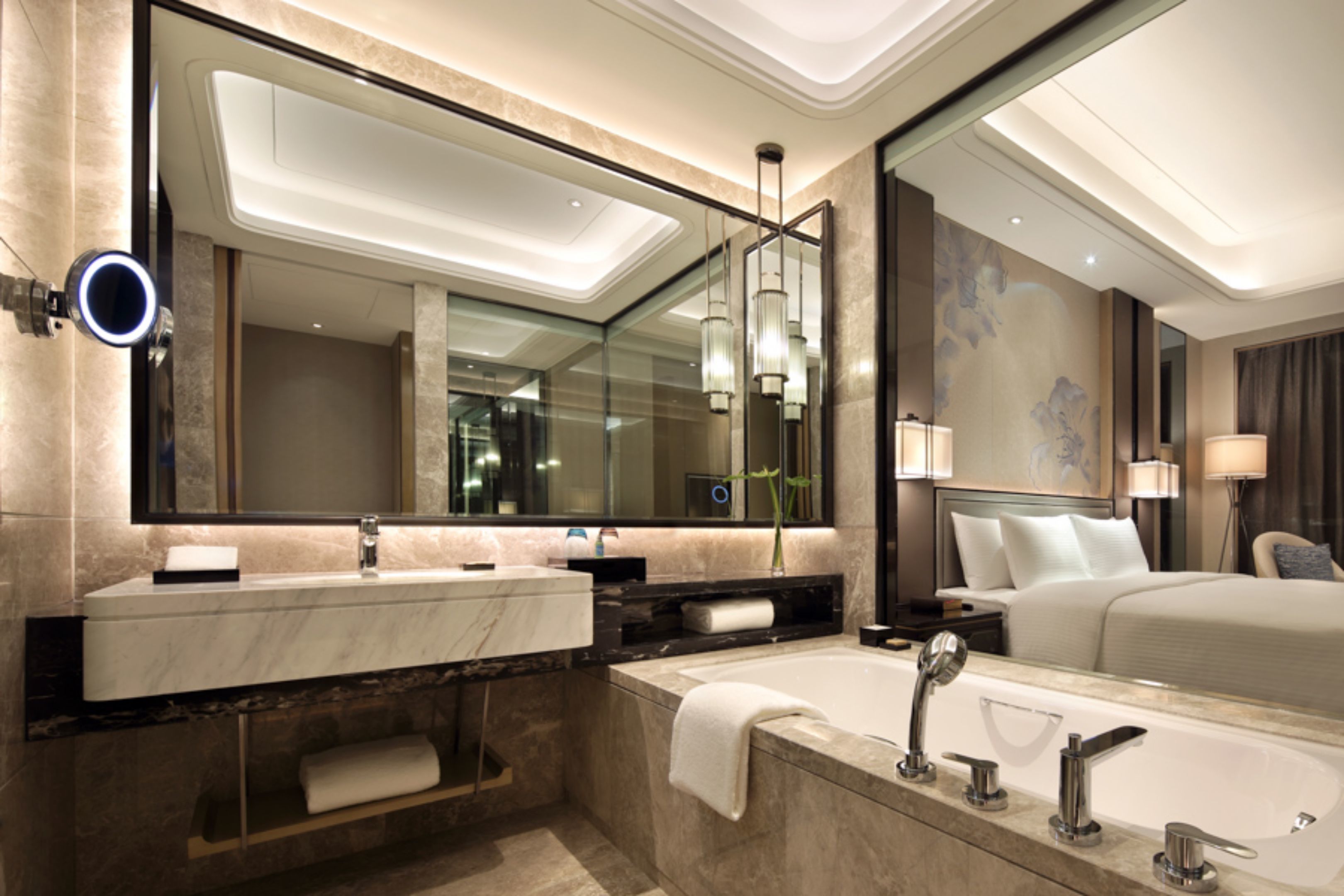 A modern hotel bathroom with a large mirror, two sinks, a bathtub, and a large bed.