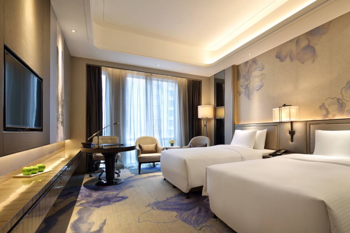 A hotel room with two beds, a desk, a chair, a TV, and a large window with curtains.