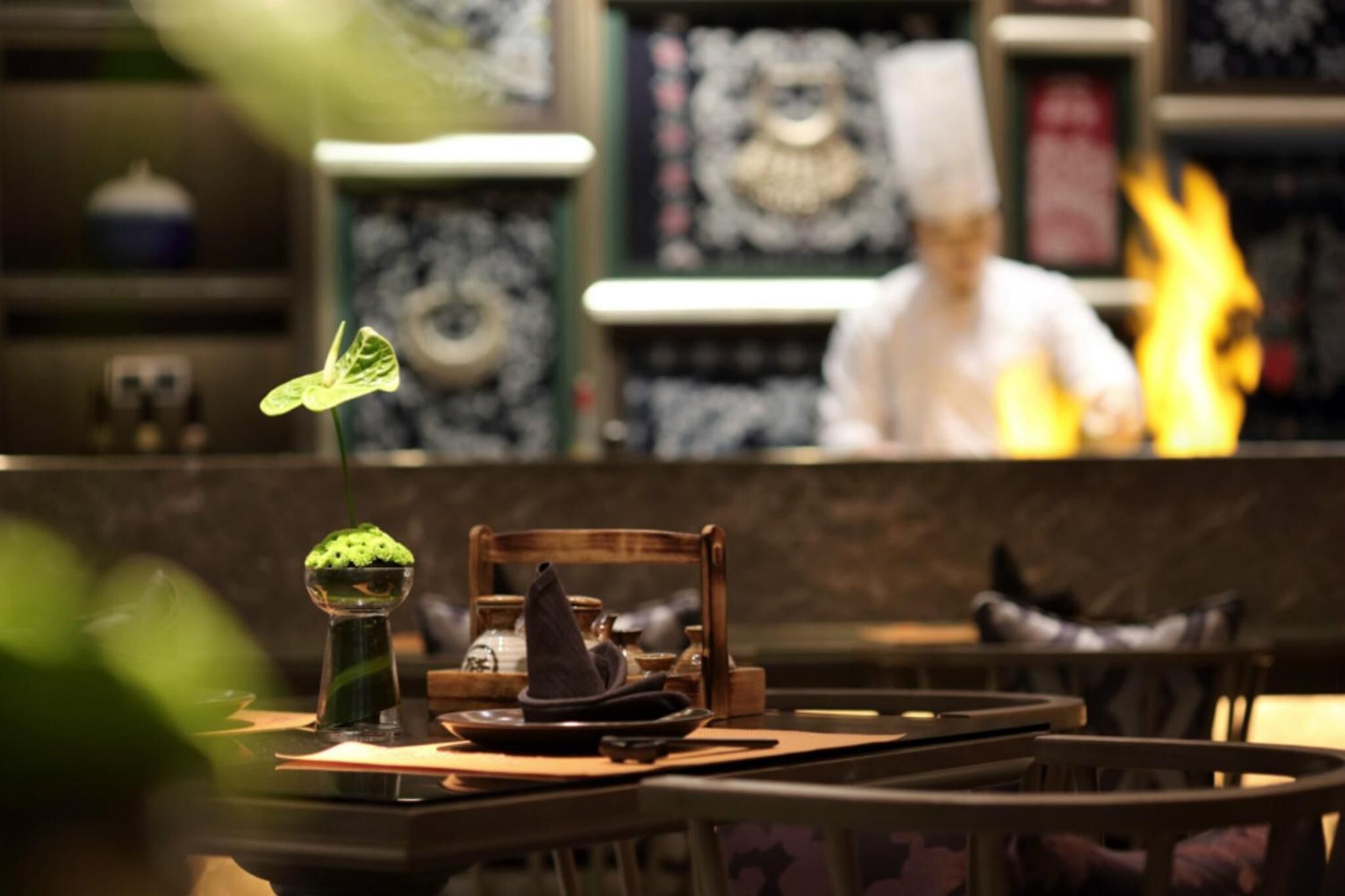 A restaurant interior with a chef in the background, a table with a green bowl and a hat, and a decorative wall with a framed picture.