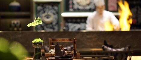 A restaurant interior with a chef in the background, a table with a green bowl and a hat, and a decorative wall with a framed picture.