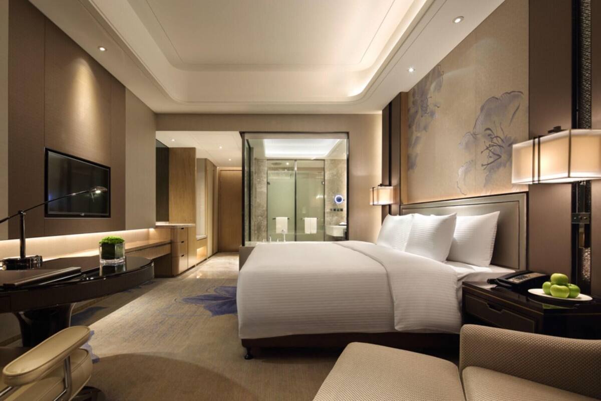 A hotel room with a bed, a desk, a chair, a TV, and a bathroom visible through an open door.