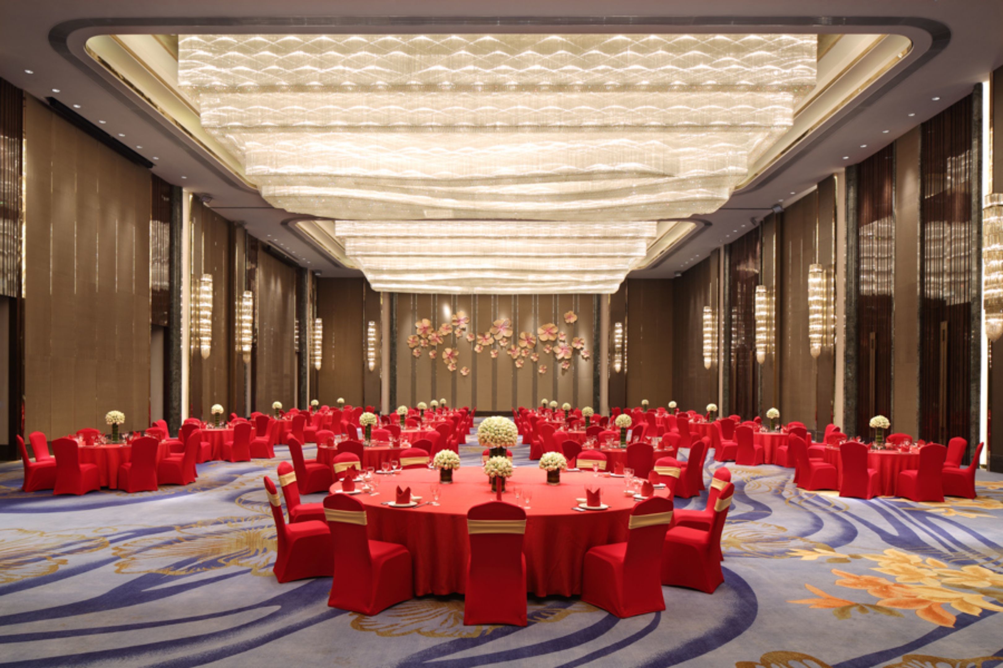 A large conference room with round tables and red chairs, featuring a high ceiling with a light fixture and floral wall decorations.