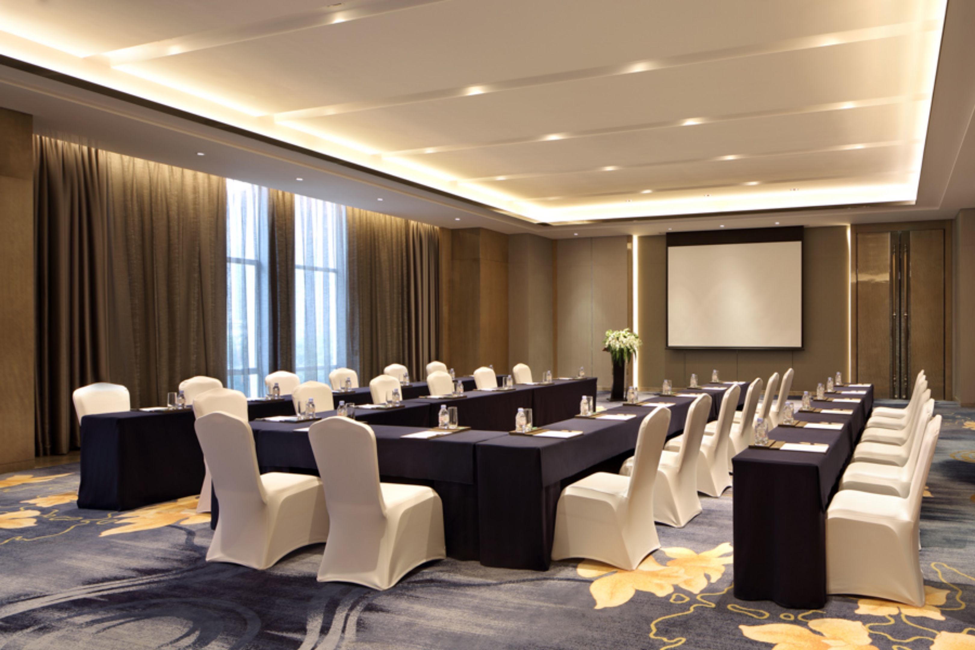 A conference room with a long table, white chairs, and a projector screen.