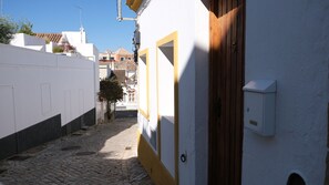 Une ruelle étroite pavée de pierres, bordée d’un bâtiment blanc avec une porte en bois et une petite boîte aux lettres.