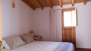 Een slaapkamer met een houten plafond, een bed met een bedsprei met patroon, een nachtkastje met een lamp en een raam met gordijnen.