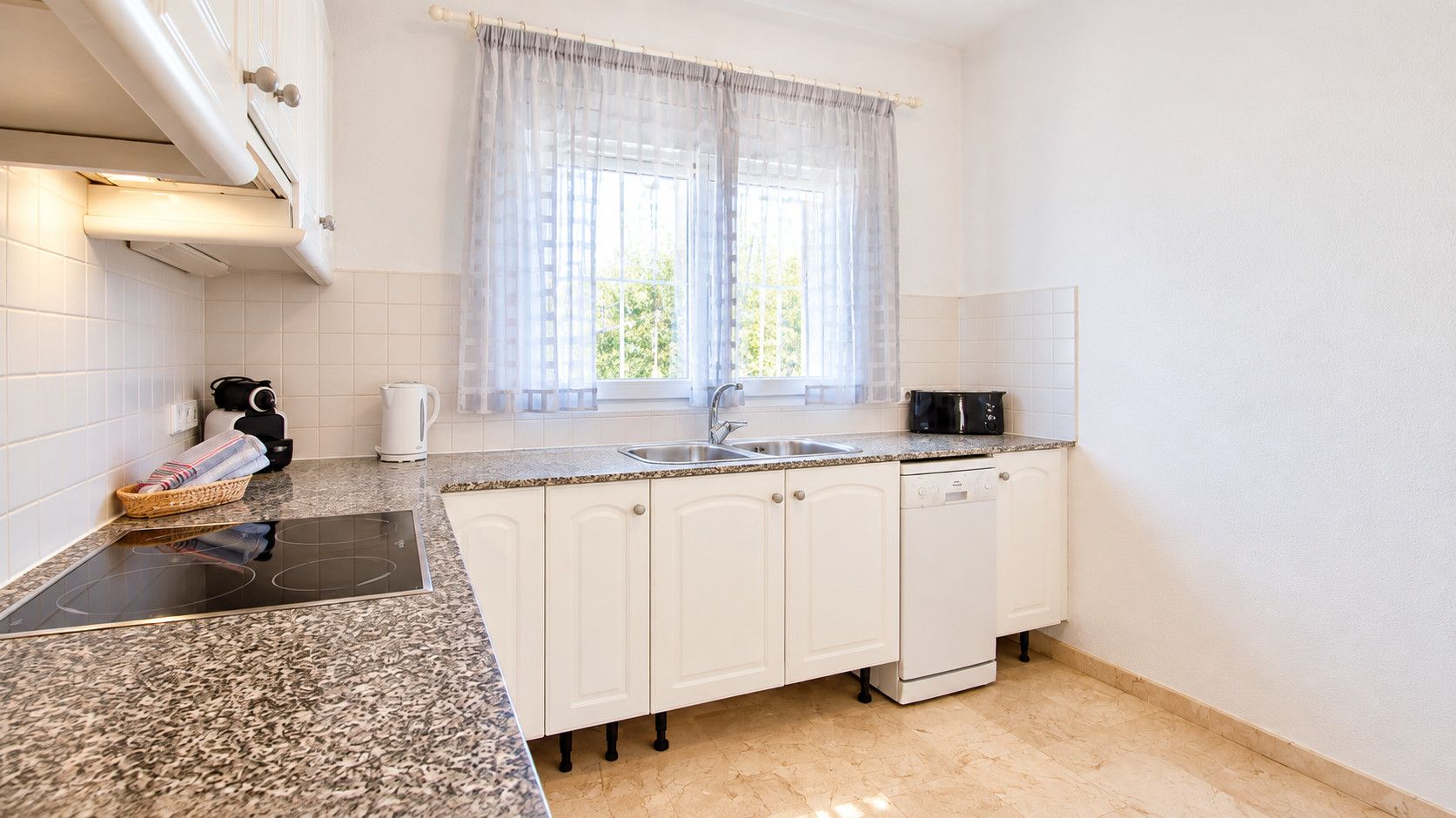 Une cuisine avec des meubles blancs, un plan de travail en granit, une cuisinière noire et une fenêtre ornée de rideaux légers.