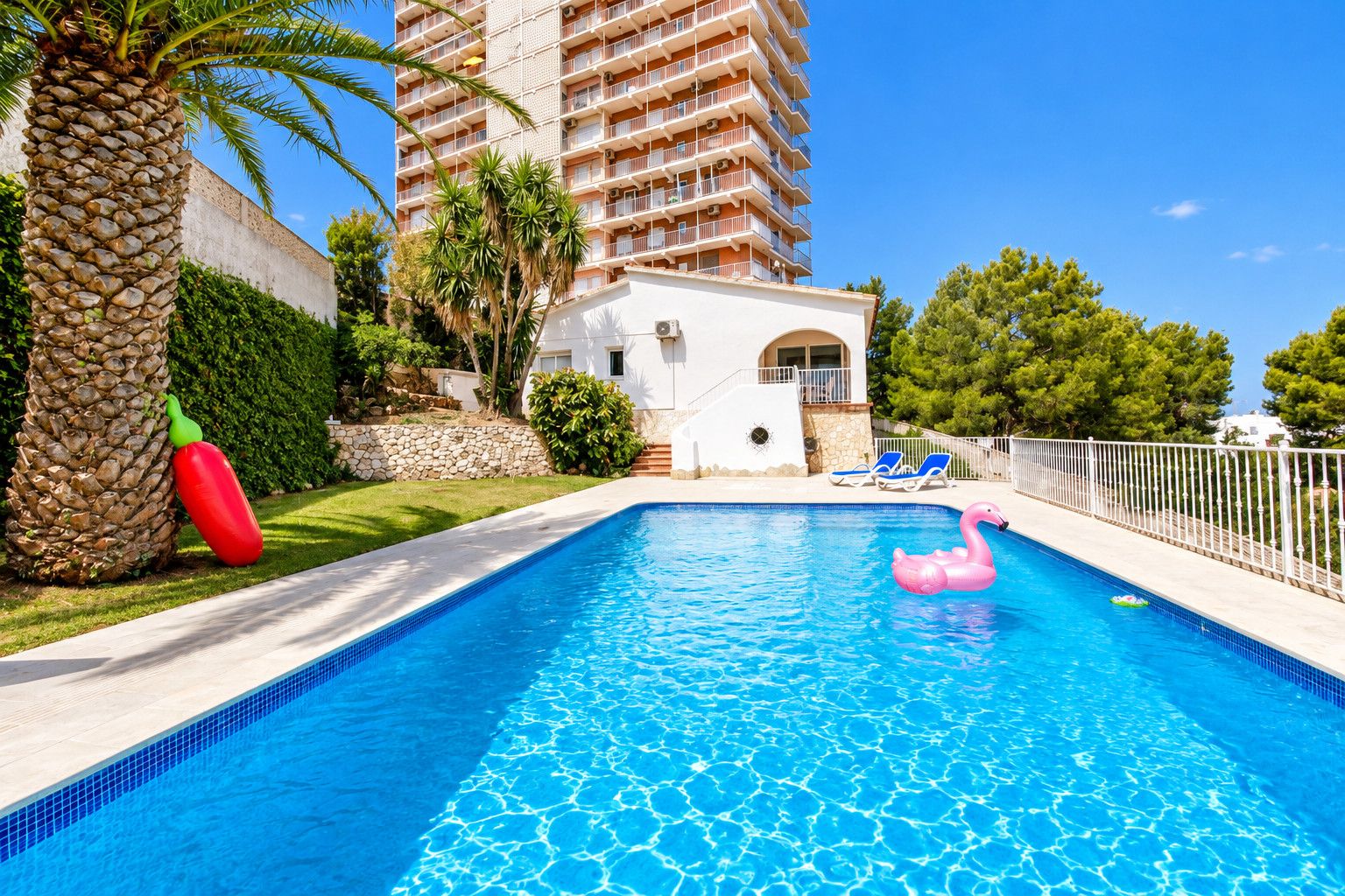 A swimming pool with a clear blue water surface, surrounded by a white fence, palm trees, and a building with balconies in the background.