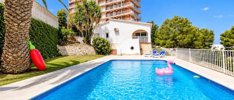 A swimming pool with a clear blue water surface, surrounded by a white fence, palm trees, and a building with balconies in the background.
