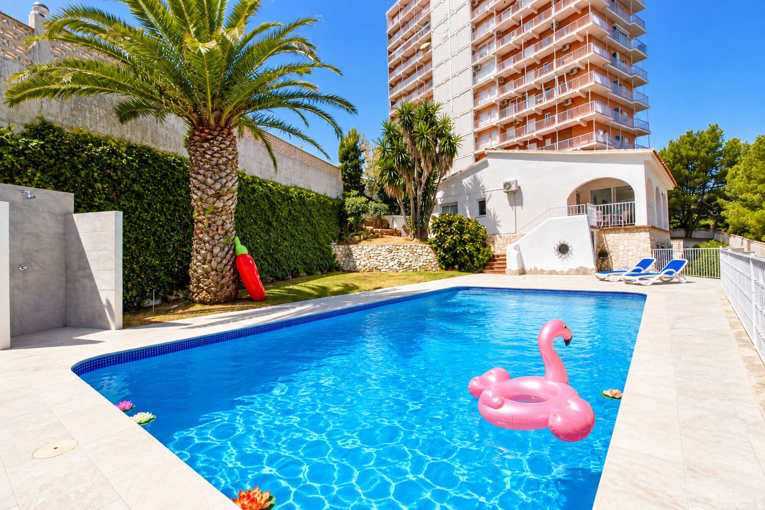 A swimming pool with a pink flamingo float, a palm tree, and a tall building in the background.