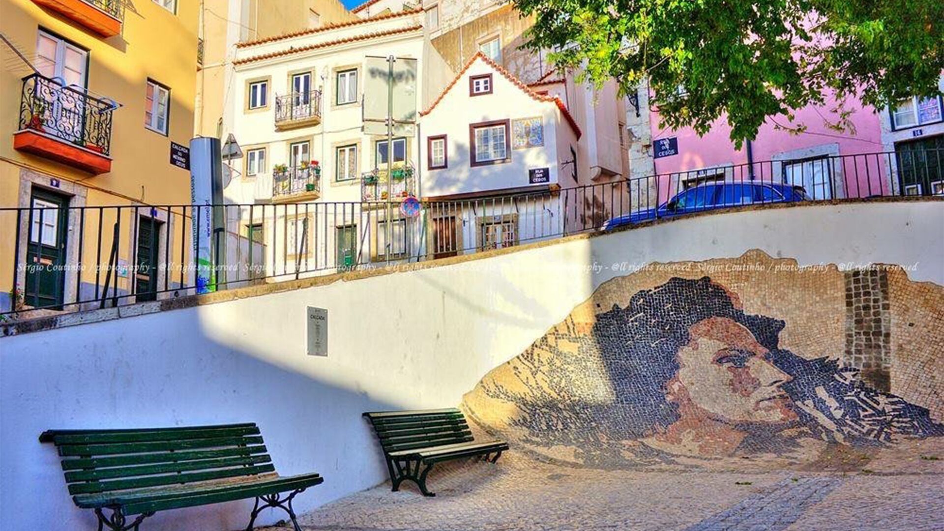 A street with benches, a mosaic wall, and buildings with balconies.