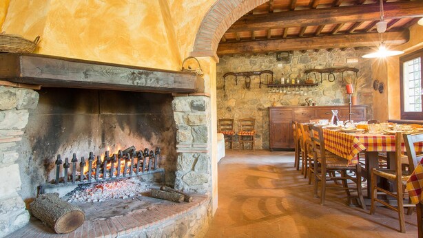 Un intérieur rustique avec une cheminée en pierre, des poutres en bois et un coin repas agrémenté d’une nappe à carreaux.