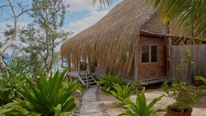 A rustic wooden cabin with a thatched roof, surrounded by lush greenery and a sandy path leading to the entrance. - Casa Algodoal (Tofo)