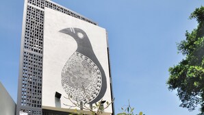 A modern building with a large bird mural on the facade, a red door, and a small tree in the courtyard.