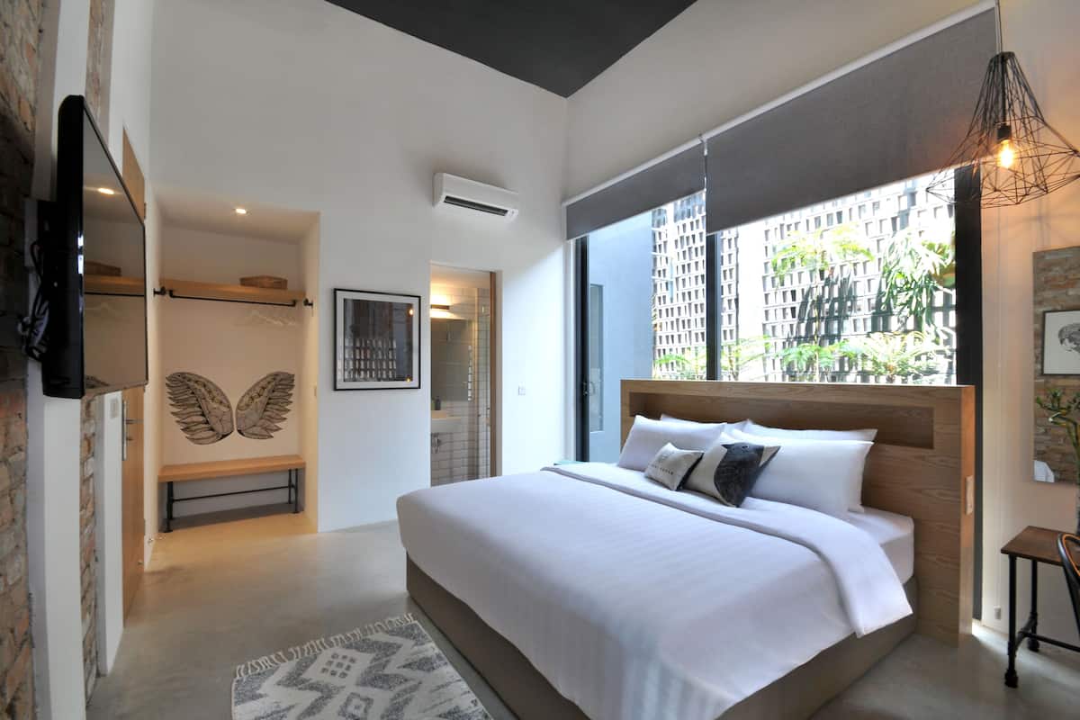 Un dormitorio moderno con una cama grande, un banco, un televisor montado en la pared y vistas a un paisaje urbano a través de un ventanal amplio.
