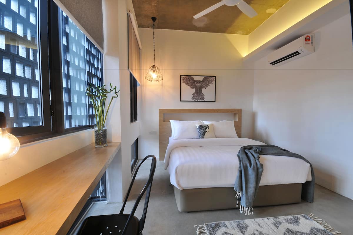 Una habitación de hotel moderna con un ventanal grande, un escritorio de madera, una cama con una manta gris y un aire acondicionado instalado en la pared.