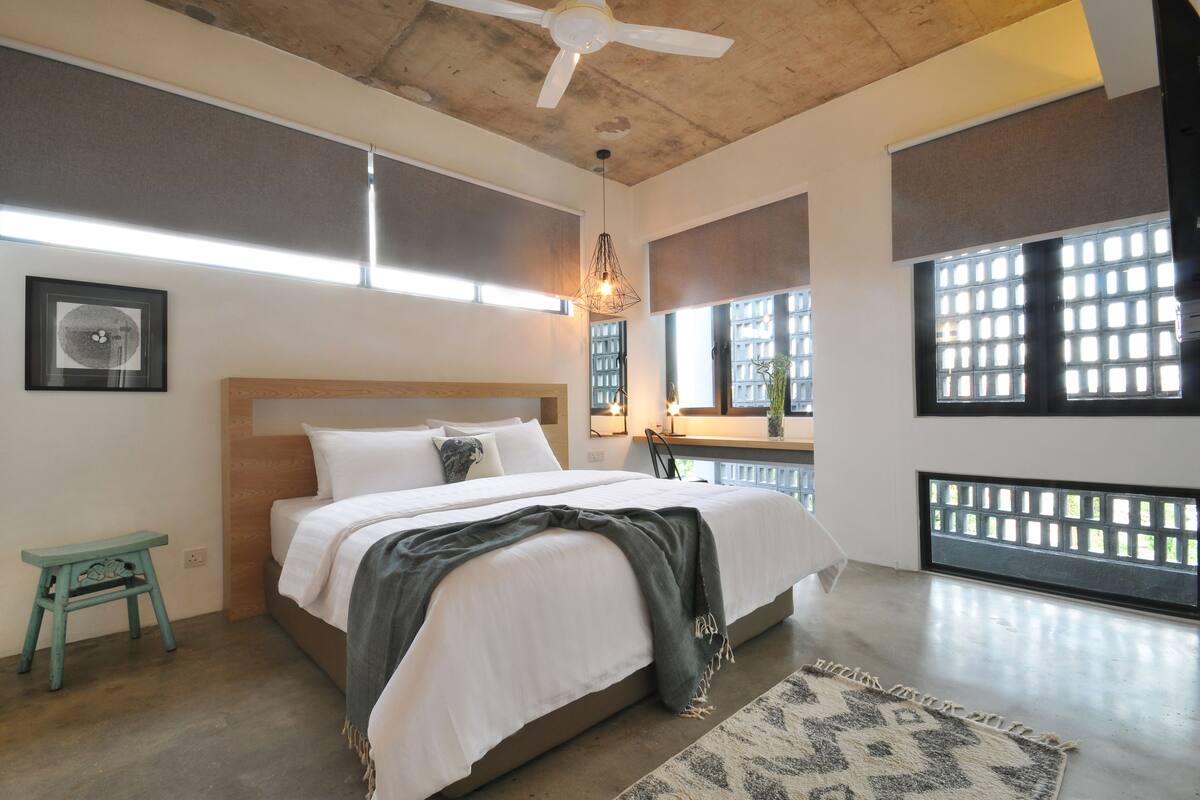 Un dormitorio con una cama, ventilador de techo, lámpara colgante, ventana con vistas y un taburete pequeño.
