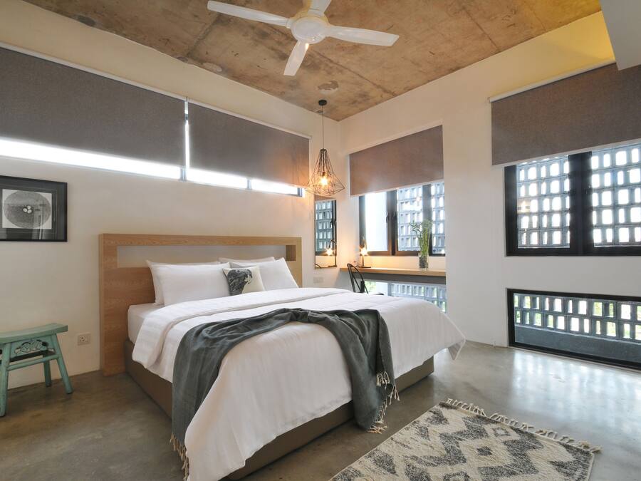 Un dormitorio con una cama, ventilador de techo, lámpara colgante, ventana con vistas y un taburete pequeño.