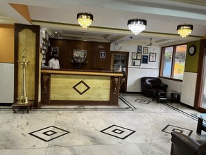 Un área de recepción con mostrador de madera, candelabros, un reloj, certificados enmarcados y piso de mármol con diseños geométricos.