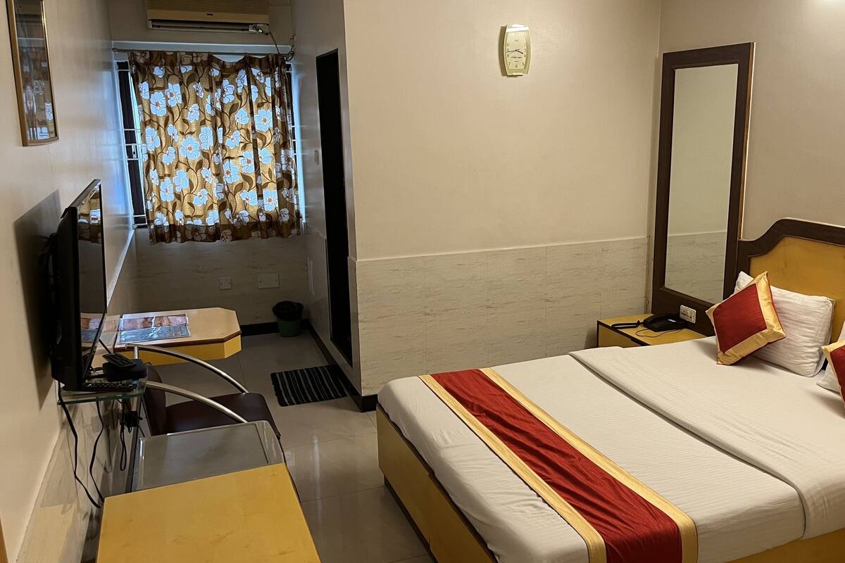 Habitació d'hotel amb un llit, una taula de treball amb un ordinador portàtil, una cadira, una televisió, un mirall i una finestra amb cortines.