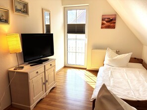 A bedroom with a TV on a dresser, a bed with white bedding, a window with blinds, and a radiator.