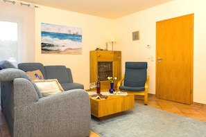 Un salon avec deux canapés gris, une table basse en bois, un fauteuil bleu, un meuble en bois et une porte.