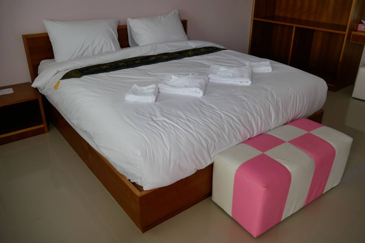 A neatly made bed with white linens and towels, a wooden headboard, and a pink and white ottoman.