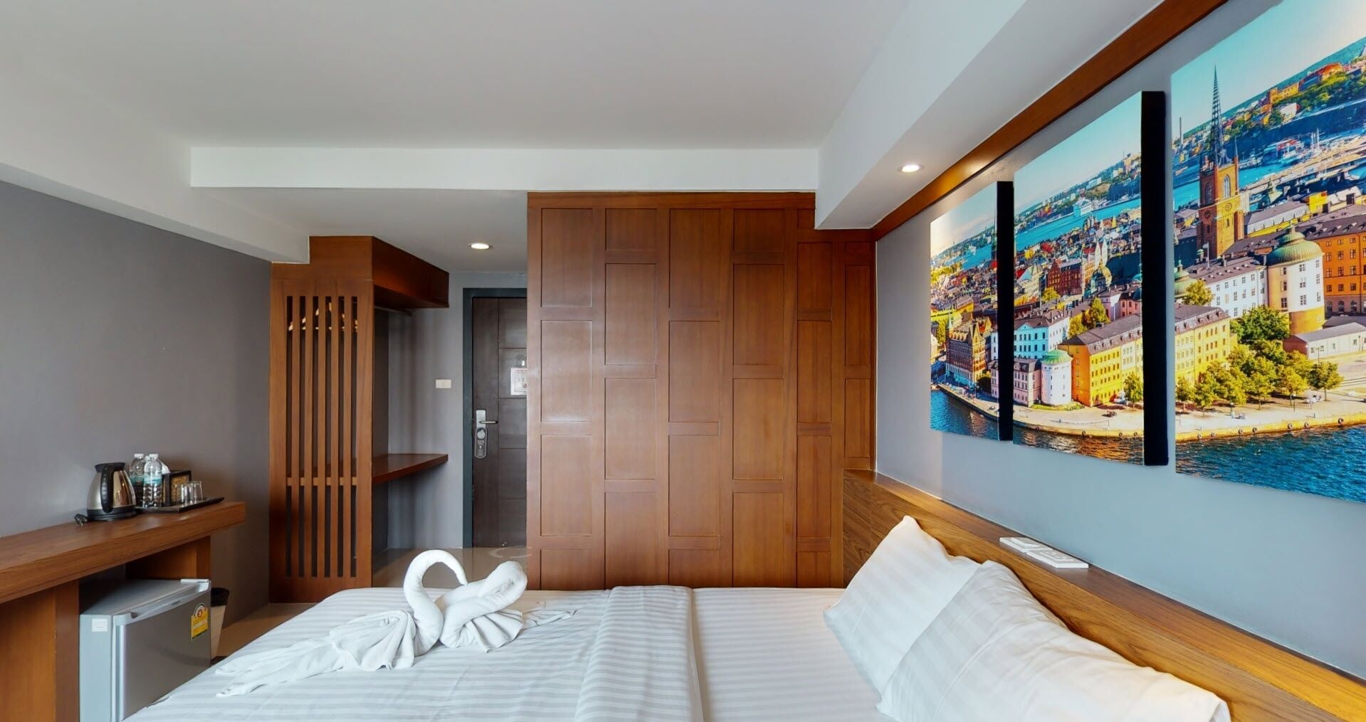 A hotel room with a bed, a wooden headboard, a painting of a cityscape, and a minibar.