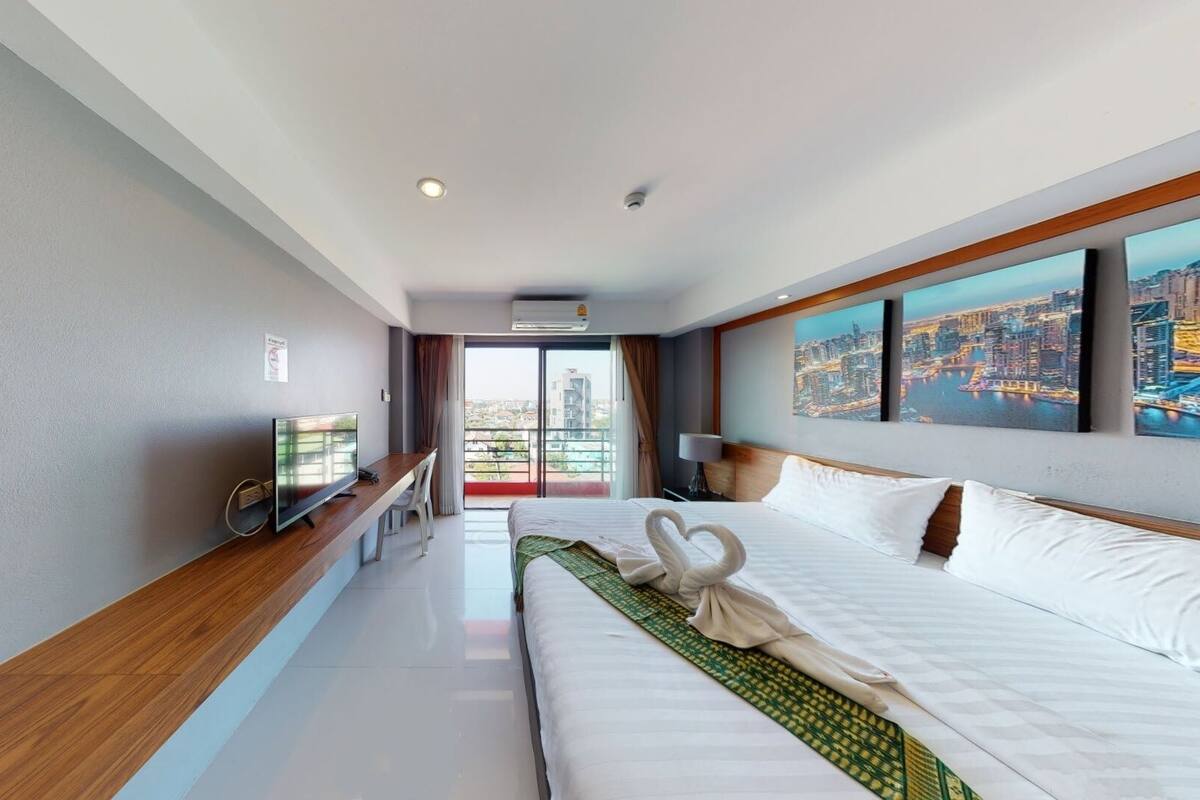 A hotel room with a bed, a desk with a TV, a balcony with a city view, and artwork on the wall.