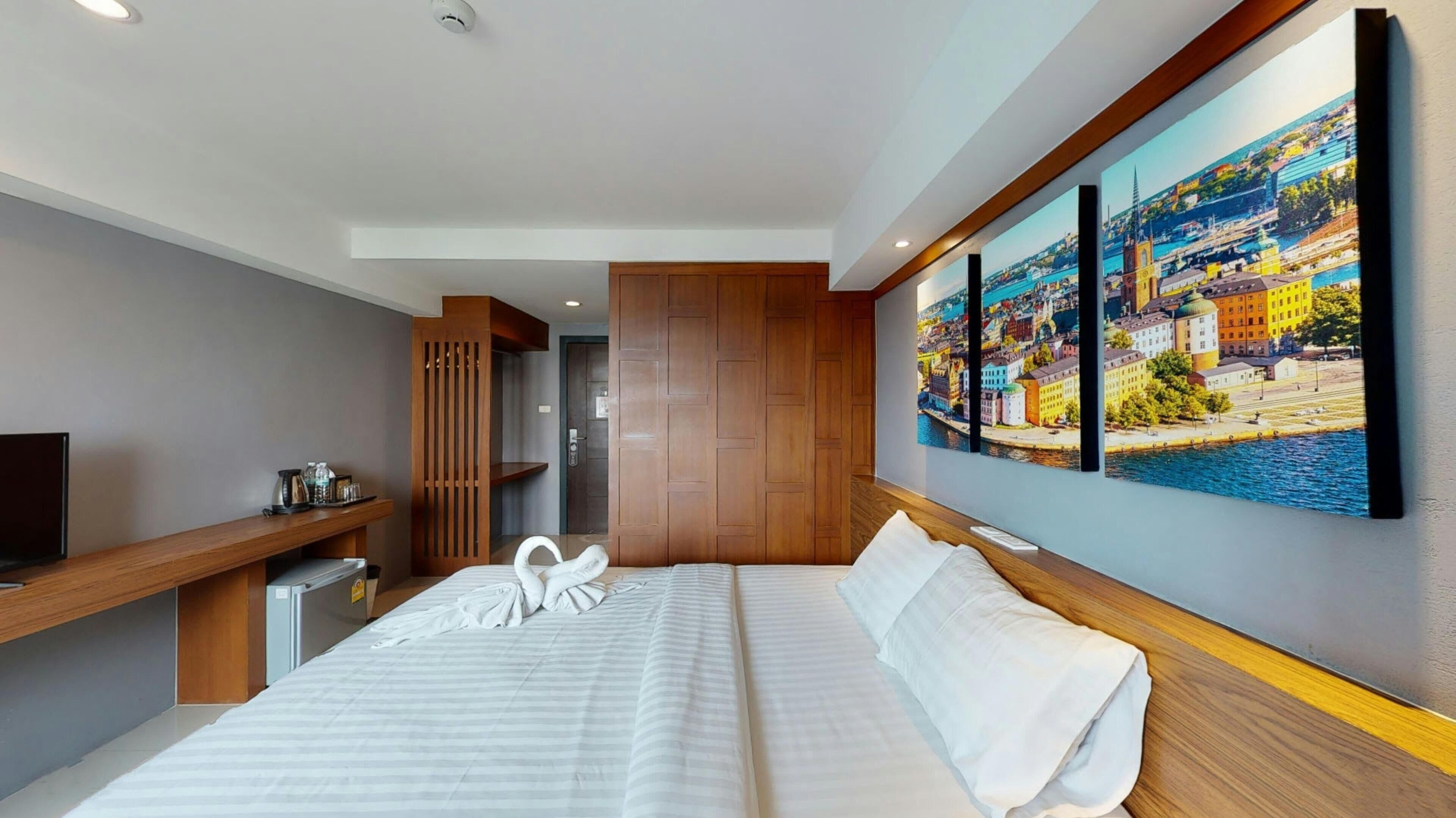 A modern hotel room with a large bed, a wooden headboard, a desk with a computer, a TV, and a large cityscape painting on the wall.